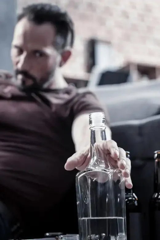 photo of an angry man sitting on the floor and reaching for the bottle while being addicted to alcohol