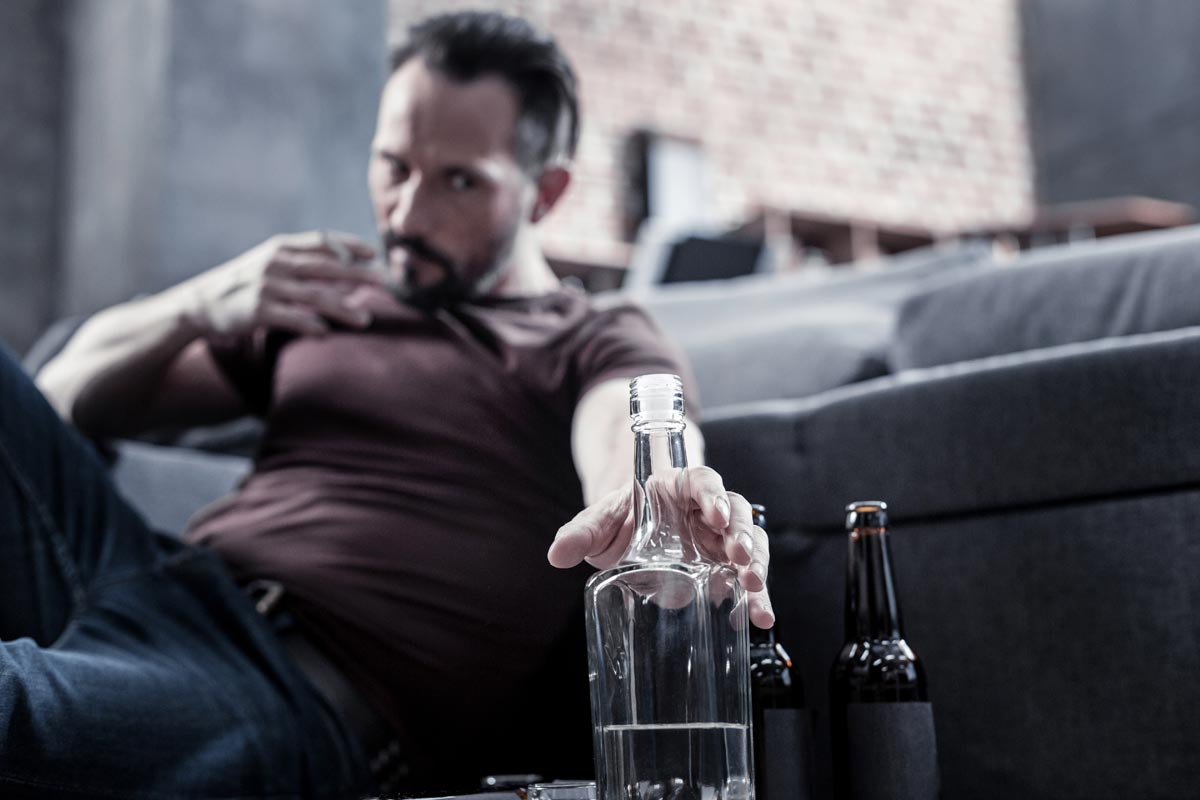 photo of an angry man sitting on the floor and reaching for the bottle while being addicted to alcohol
