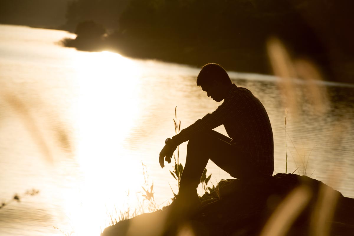 young man with depression sitting near a river during sunset silhouette