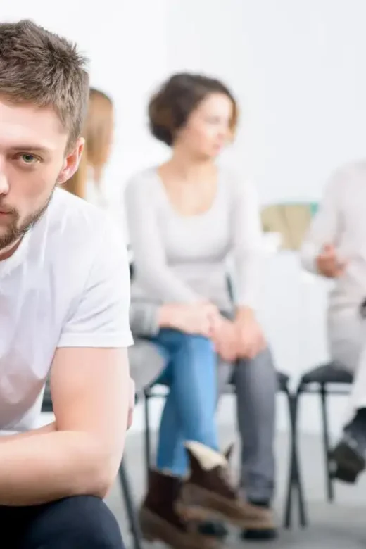 Depressed cheerless boy sitting in the chair with professional psychologist working in the background with people during psychological recovery group therapy session