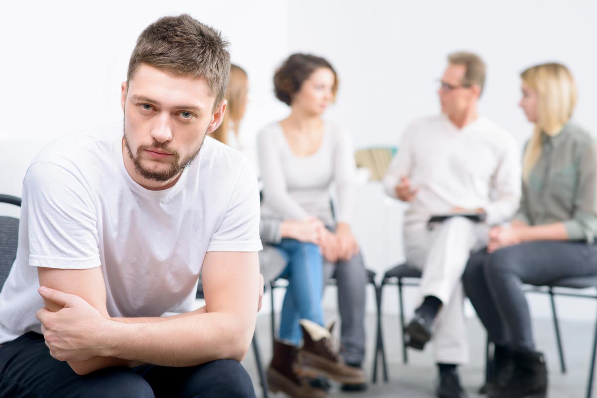 Depressed cheerless boy sitting in the chair with professional psychologist working in the background with people during psychological recovery group therapy session
