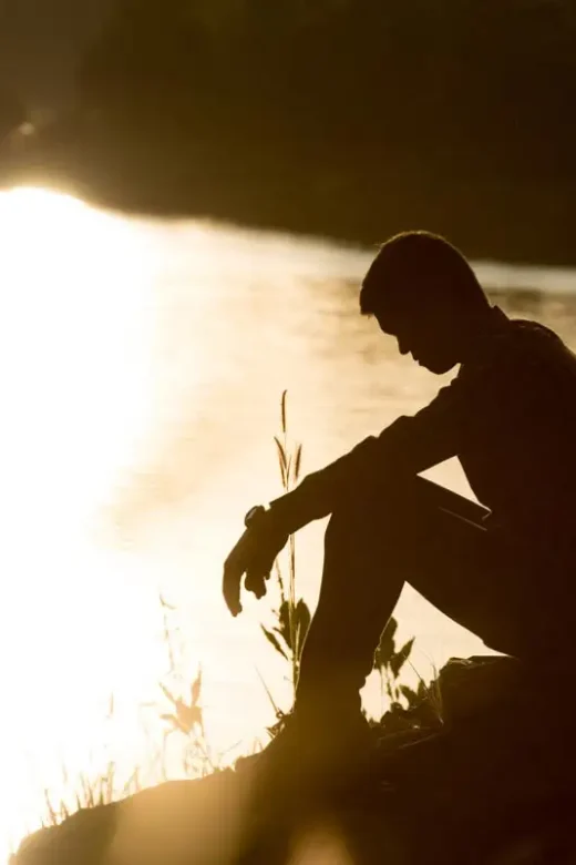young man with depression sitting near a river during sunset silhouette