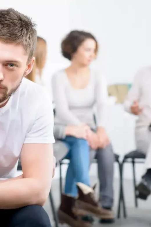 Depressed cheerless boy sitting in the chair with professional psychologist working in the background with people during psychological recovery group therapy session