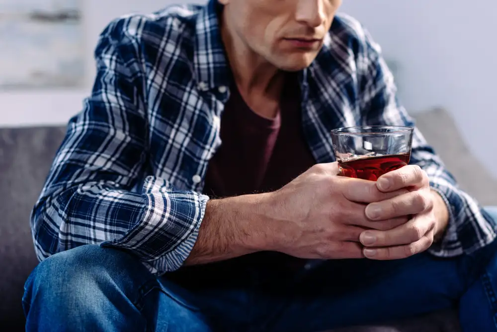 A man sits on a couch, casually holding a glass of hard liquor suffering from alcohol abuse