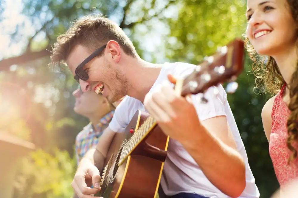 two men and a woman sit on a bench at Riverside Inpatient Rehab, playing an acoustic guitar together, enjoying their time.