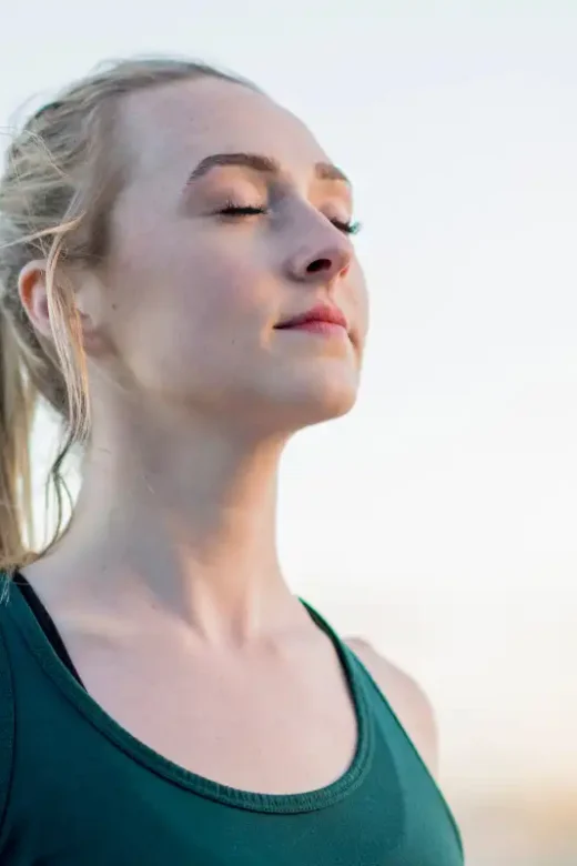 Beautiful blonde young woman wearing a green fitness shirt has her eyes closed and chin up in meditation while performing yoga outdoors in a public park.
