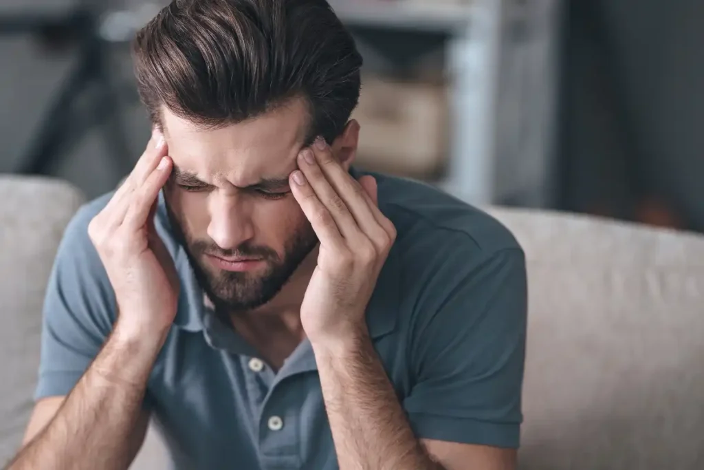 Frustrated handsome young man touching his head and keeping eyes closed while sitting on the couch at home