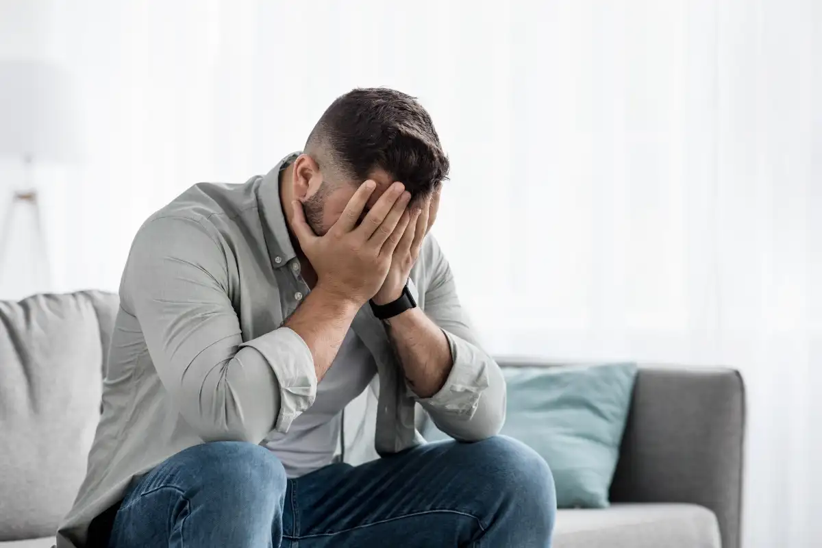 unhappy guy sitting on a sofa, disappointed covering his face with hands