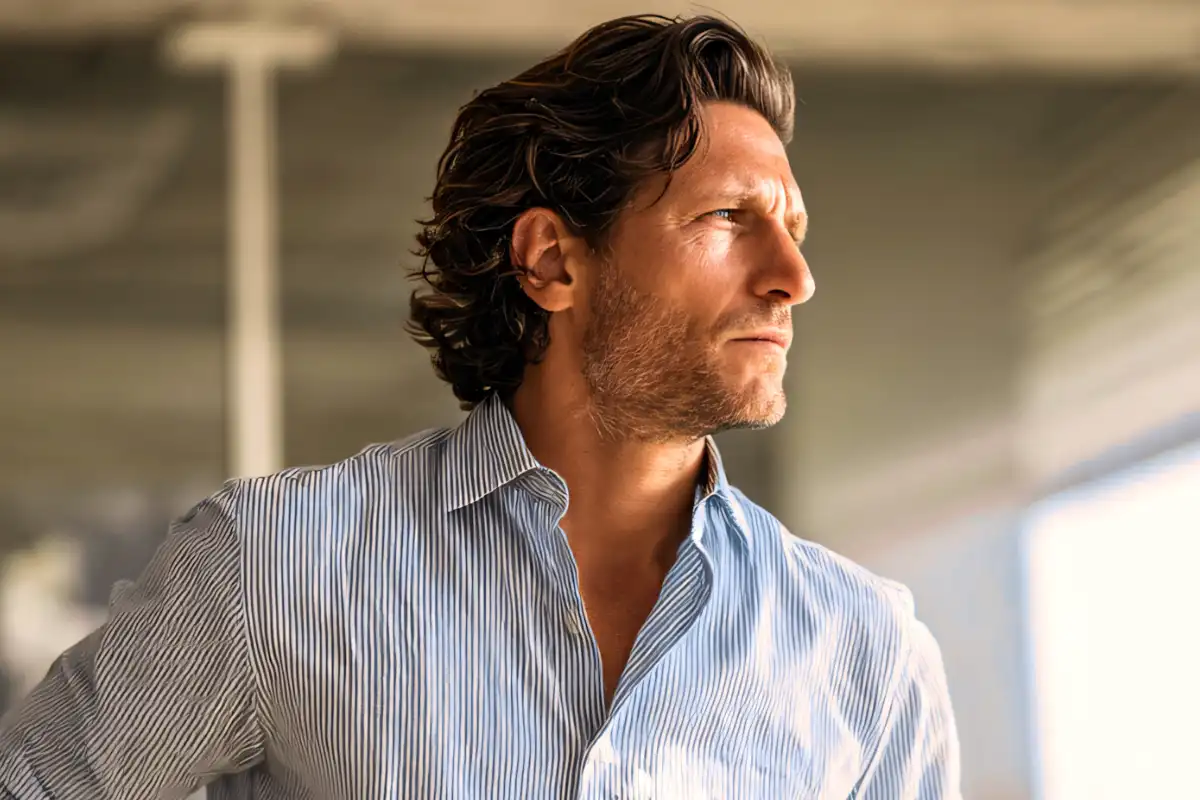 A man with curly brown hair, wearing a light-colored striped shirt and beige pants, stands with his hands in his pockets, looking thoughtfully out a window.